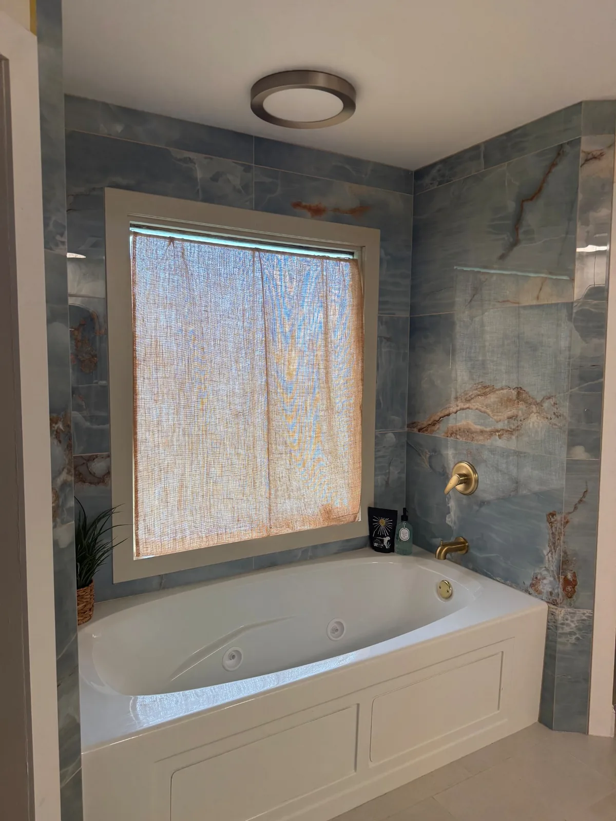 White bathtub under a window, surrounded by blue stone-look tile and brass fixtures
