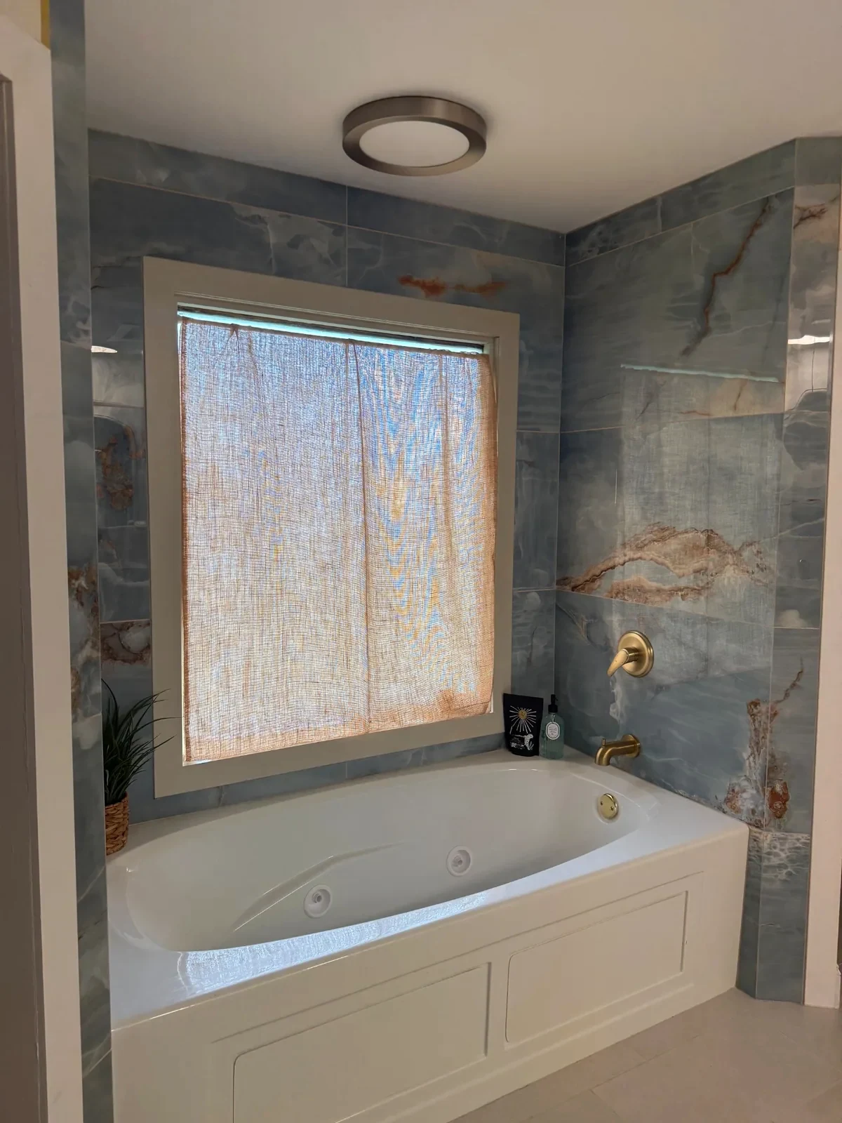 White bathtub under a window, surrounded by blue stone-look tile and brass fixtures